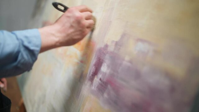 Creative person at work, adult man talented artist covers the canvas with colorful oil paint, priming and draws the background, close-up view of hands.