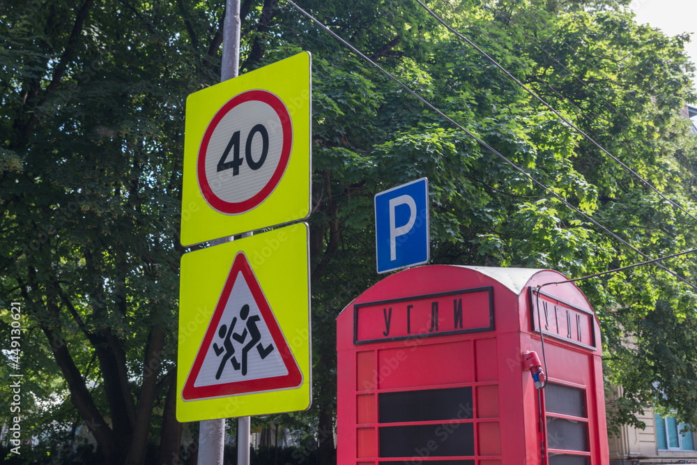 Wall mural Road signs Caution, children and Speed limit 40 on pole with wires, next to them red booth with inscription in russian COALS, behind it blue parking sign against background of dense foliage of trees.