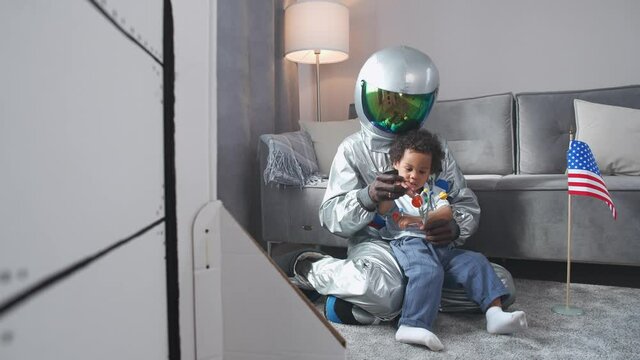 Afro american father and son play in the living room at home, man in a suit of an american astronaut sitting on the floor with her son, boy playing with a toy model of the solar system, study of