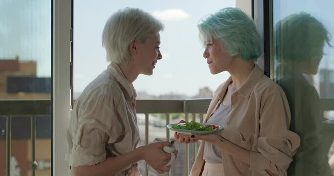 Side View On Two Attractive Women In Domestic Clothes Treating Each Other With Breakfast On Balcony, Smiling, Enjoying Morning Routine Together. Copy Space. Leisure, Love, People Relationships, Lgbt