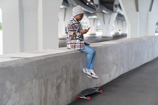Trendy african skateboarder girl with smartphone in hands sitting on concrete pavement read chat message from friend in mobile phone. Longboarder female with digital gadget outdoors scrolling browser