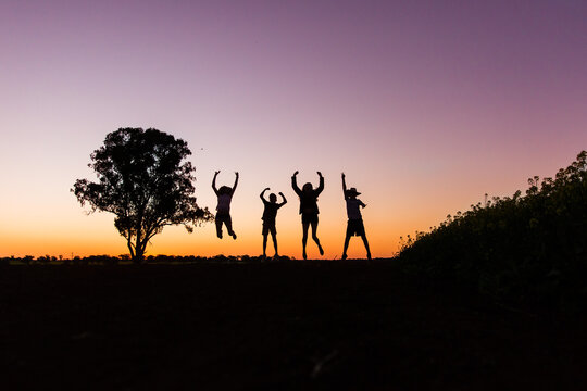 Silhouette Of Four Children Jumping Near Tree In Paddock On Farm At Sunset