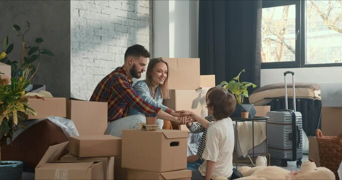 Young happy family with two children sit on couch in apartment with boxes after moving in, join hands to celebrate.