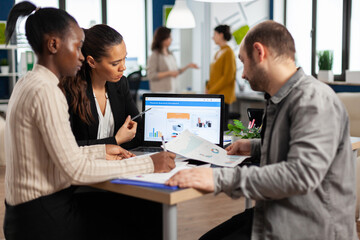 Busy multicultural, diverse employees analysing annual financial statistics sitting at desk in front of laptop holding documents searching business solutions. Team of businesspeople working in company