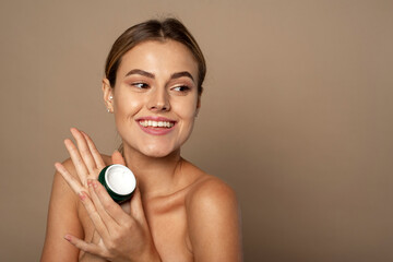 Cute woman holding a jar of moisturizer in the studio. Skin care and hydration concept. Portrait of beautiful woman applying some cream to her face for skin care