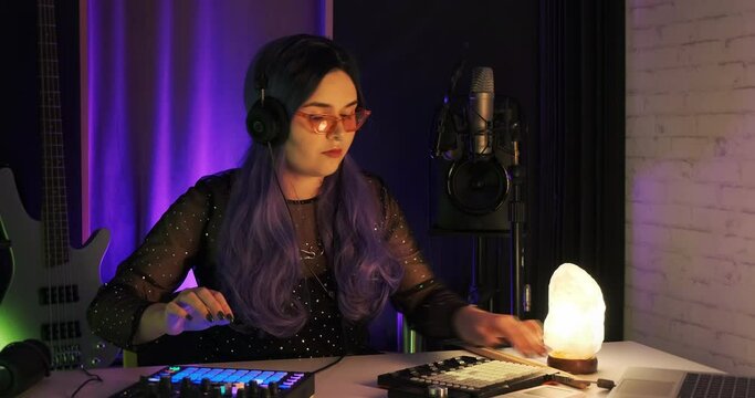 Medium shot of female LGBTQ non-binary Dj musician writing beats on push controller. Bas guitar and ambient lighting in small bedroom studio. Music rehearsal session, colored hair, alt girl tik toker
