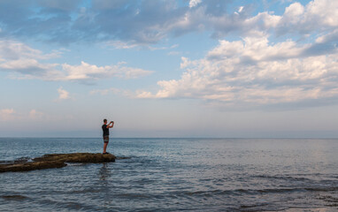 Young caucasian man in shorts taking photo with mobile phone on Croatia sea rock shore edge under cloudy sky. Side view, copy space