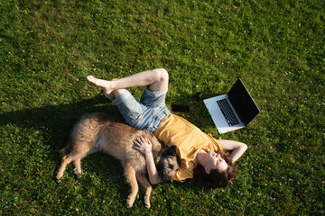Beautiful happy young woman lying on the grass with laptop and dog, top shot