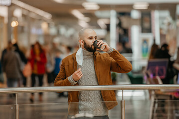 A man is holding a took-off mask while drinking coffee in the shopping center.