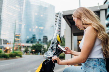 Young caucasian woman outdoors sitting sharable electric scooter outdoor unlocking using smartphone
