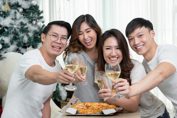 Group of Asian young male and female friends gather to celebrate Christmas, clinking champagne glasses and eating pizza at home. Joy of holiday party with friends or family concept