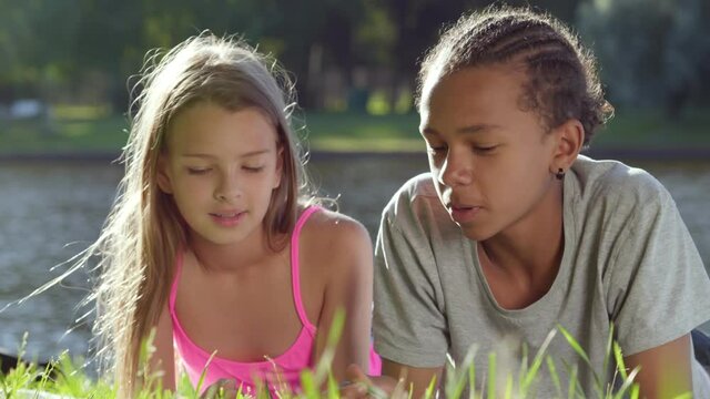 Caucasian teen girl tell secret to african boy lying together on grass in park