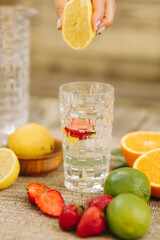 A tall glass cup of water with ice. Sliced oranges, lemons, lime and strawberry. Woman squeezes lemon juice in water