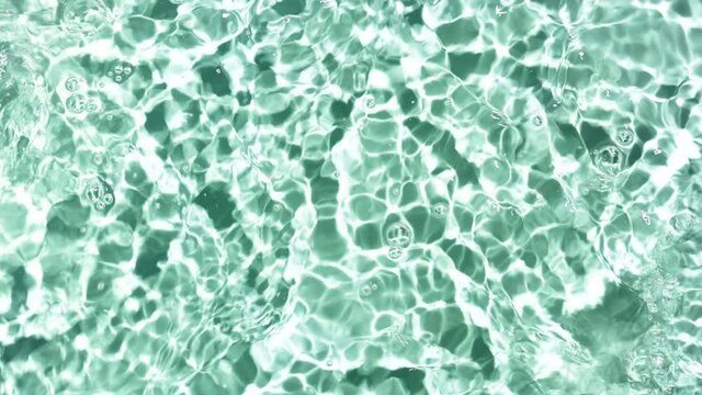 Multiple waves of water cross each other and a lot of drops fall down on water surface creating ripples and bubbles spontaneously on pale green background | Cosmetics background shot for advertising