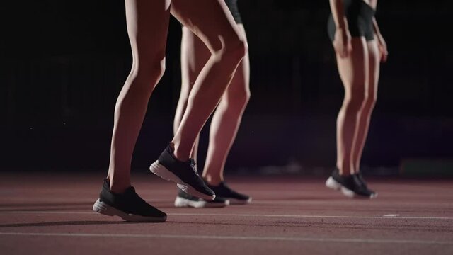 Three women athletes prepare for a track race in a dark stadium with streetlights on. Time-lapse footage of warm-up and concentration of a group of women before the race on the track
