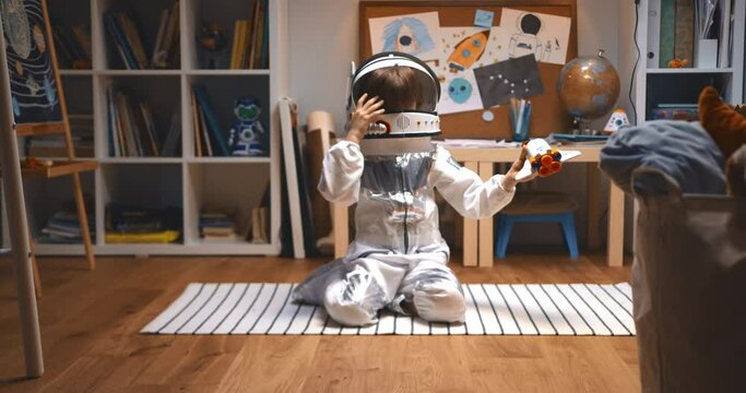 Childhood dreams about future. Happy little boy in astronaut suit playing space ship pilot, flying rocket toy on floor.