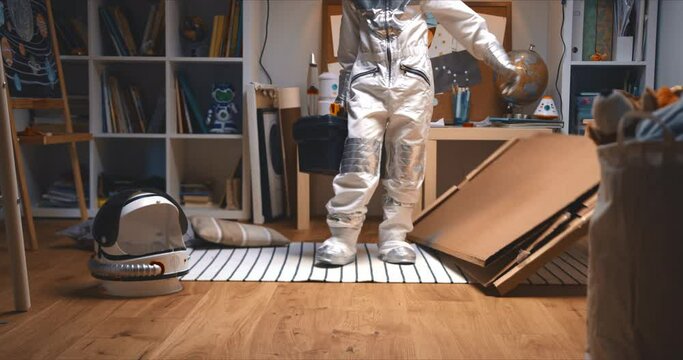 Childhood activities. Little boy in astronaut suit enters room with big cardboard piece, tool box to build space ship.