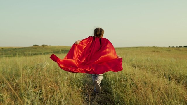 Child plays and dreams. Happy superhero girl, runs on green field in red cloak, cloak flutters in wind. Teenager dreams of becoming superhero. Young girl in red cloak, dream expression.