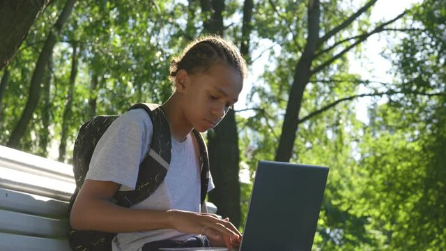 Afro-american teenage boy with laptop sitting on bench in park.