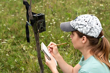Young woman scientist zoologist writing down data from trap camera to notepad, observing wild...