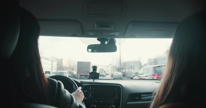 View from backseat, two beautiful happy female friends in face masks talk while driving car along busy city streets.