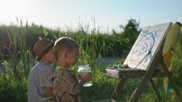 lifestyle, little boy drinks water and draws together with his brother with brushes and paint on paper while relaxing in nature on background of green grass and reeds