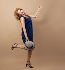 Smiling woman with blond hair, dressed in a blue sundress, dancing with a hat in her hand, isolated...
