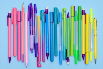 Rainbow stationery collection of pens, ballpoints and pencils on a light blue background. Lots of pink, purple, blue, green and yellow office and school supplies in a row.