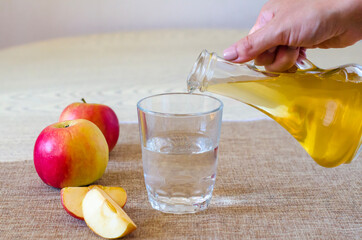 A woman's hand pours apple cider vinegar in a glass bottle on a light background into a glass of...