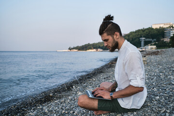 Naklejka na ściany i meble Successful young Caucasian male freelancer is sitting on beach and remotely working online using laptop. Hipster businessman works on travel and runs blog on the Internet. Focused job outside.