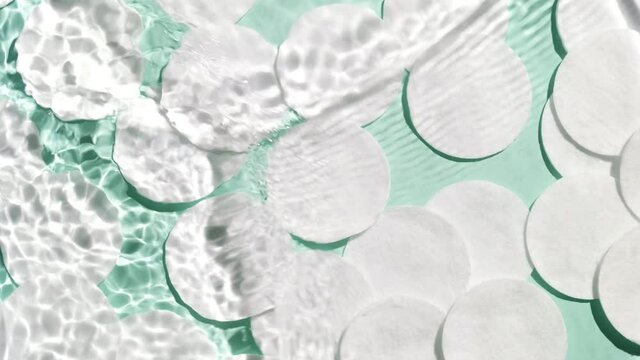 Multiple waves of water cross each other creating ripples spontaneously on pale green background with cotton pads | Background shot for face care cosmetics commercial