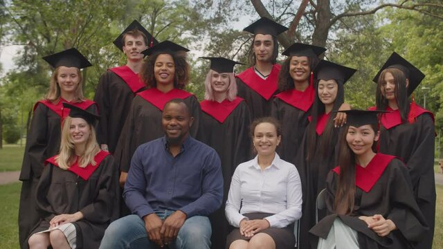 Closeup of carefree multiracial diverse graduates in gowns and mortarboards with cheerful multiethnic professors posing for picture together, showing positive vibes after receiving university diploma.
