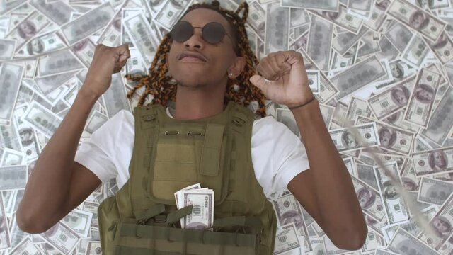 African-American man in bulletproof vest is lying on pile money and dancing, money rain. Black rapper is lying in pile of dollars bills are falling on it. Concept rich