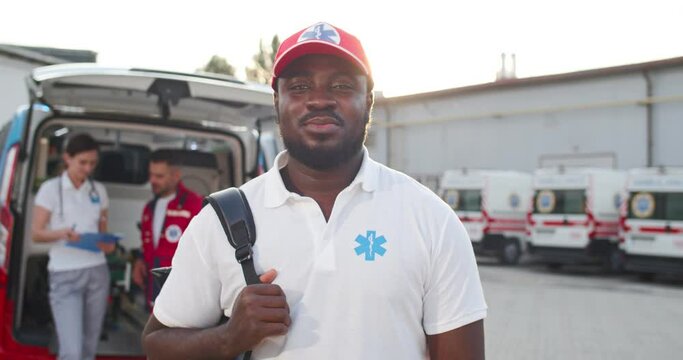 Portrait of young African American cheerful man medic in cap smiling to camera outdoors. Ambulance with colleagues paramedics on background. Male happy doctor with backpack is ready to work.