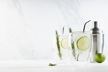 Organic mojito cocktail cooking with citrus fruit slices, ice cubes, green rosemary twig, ingredients, silver shaker and straws  in two transparent mugs on white wood table, soft light marble wall.
