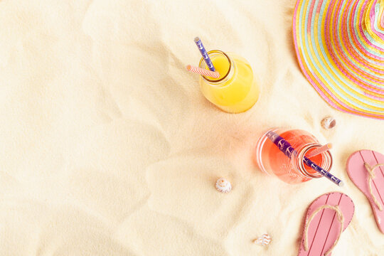Healthy summer beach vacation on ocean coast - cold pink and orange fruit juice in glass bottles, sun hat, slippers, seashells, top view, copy space.