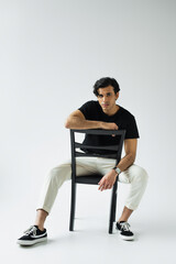 full length of young man looking at camera and sitting on chair on grey