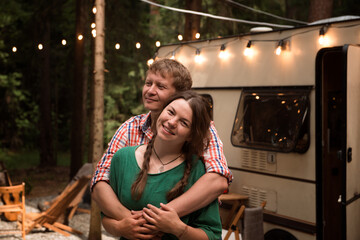 Fototapeta na wymiar Couple camping in trailer park. Happy blond man hugging pretty woman close to mobile home parked in forest. Summer vacation in tiny house on wheels