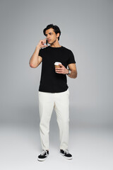 full length of young man holding coffee to go and talking on smartphone on grey