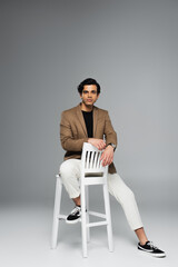 full length of young man in blazer sitting on white chair on grey