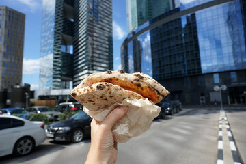 Neapolitan pizza rolled like a wallet in hand against the backdrop of modern skyscrapers.