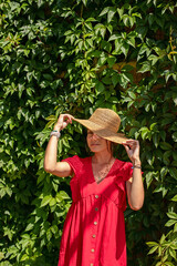 A young girl in a red dress and a straw hat poses against a green wall with ivy. Summer style.