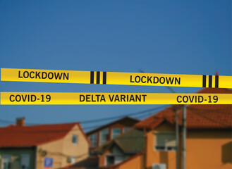 Yellow restricted area tape stripes with written text LOCKDOWN Covid-19 DELTA VARIANT against the blurred architecture of european town with red roofs and blue sky