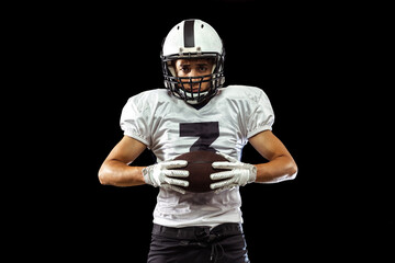 Close-up portrait of American football player in sports equipment, helmet and gloves isolated on dark studio background. Concept of sport, competition