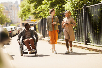 Handicapped young African-American man in glasses using manual wheelchair while walking with...