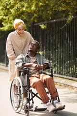 Fototapeta na wymiar Smiling young middle-eastern caregiver in hijab oushing wheelchair with disabled black man while walking with him along street