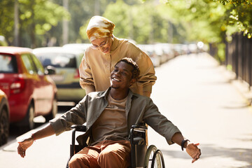 Positive young African-American disabled man chatting with friend who pushing him in wheelchair along street