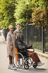 Smiling interracial friends standing by African-American man sitting in wheelchair against fence