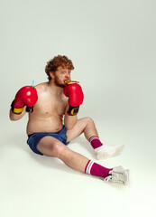 Funny red-headed man in blue boxing shorts and gloves isolated on gray studio background. Concept of sport, humor and body positive.