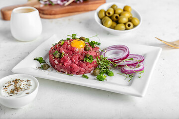 Plate with tasty beef tartare, olives and sauce on light background